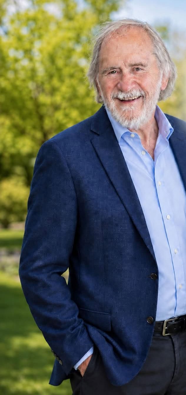 Tom Wilkins standing outdoors in a blue blazer, smiling, with green trees behind him
