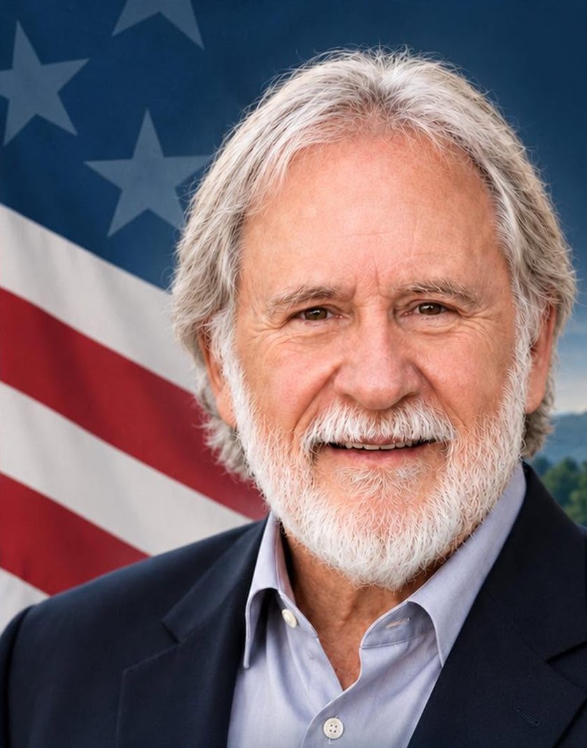 Portrait of Tom Wilkins, smiling, in a navy blazer in front of an American flag
